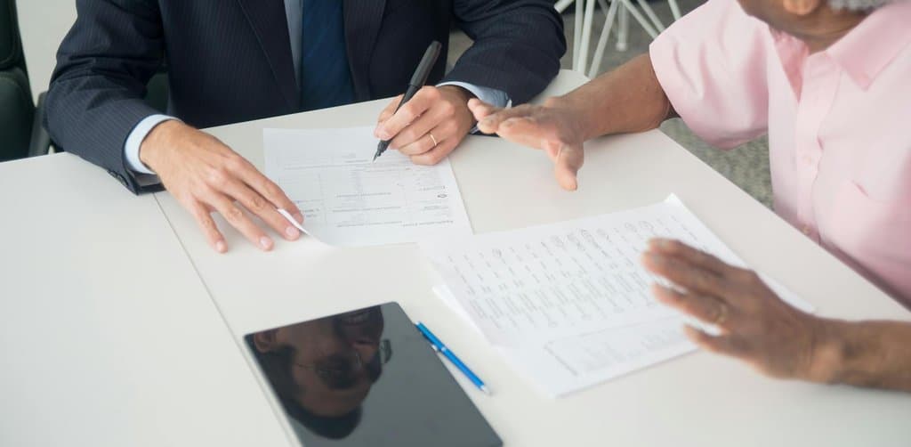A suited buyer seated across a white office table from a grey-haired seller in a pink shirt, reviewing an application form and a printed business profile, the across-the-desk conversation that defines buying a business with seller financing