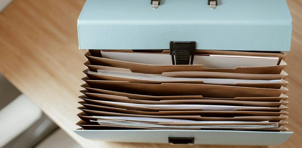 Light blue accordion file box on a wood desk holding rows of brown folder pockets stuffed with documents, the kind of deal-diligence file a buyer builds while screening small companies for sale
