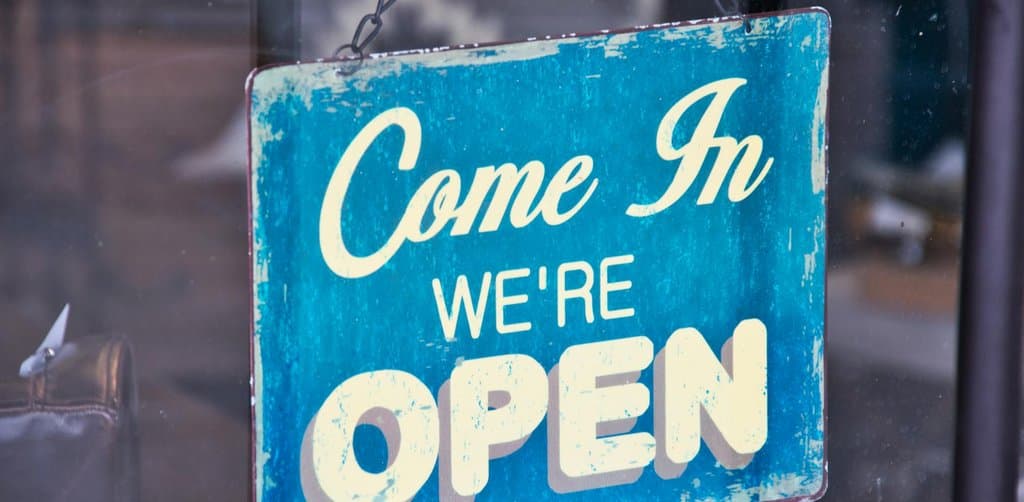 Vintage blue 'Come In We're Open' sign hanging on the inside of a small storefront glass door, the moment a new owner takes over after buying a small company and reopens the doors under new management