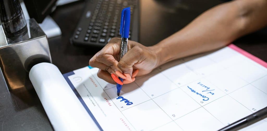 A close-up of a black womans manicured hand with orange-coral nail polish gripping a blue felt-tip Sharpie marker writing on a flip-chart paper desk calendar laid flat on a black tabletop with Friday and Thursday column headers visible the writer making notes in blue handwritten characters across the date squares with a black wireless computer keyboard and grey wireless mouse partially visible behind the calendar in soft warm office lighting, the timing-decision metaphor for the calendar windows that govern when can i sell my esop shares becomes a yes answer