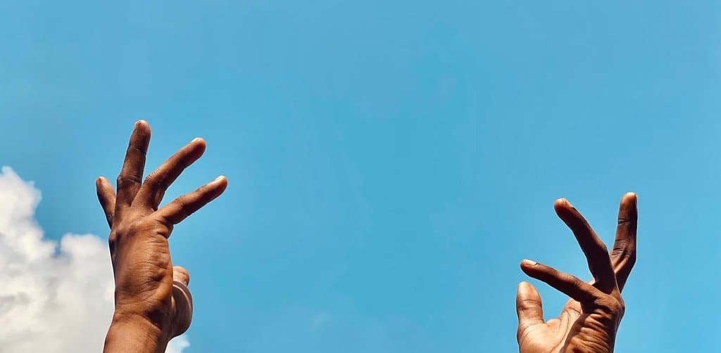 A close-up upward view of two dark-skinned hands with palms turned outward and fingers spread wide held high against a bright blue summer sky background with one fluffy white cumulus cloud at the lower left edge and another wisp of white cloud at the lower right both hands stretched up freely toward the sky in a gesture of openness or release, the literal letting-go metaphor for the moment when the answer to can i sell my esop shares becomes yes at a distribution event