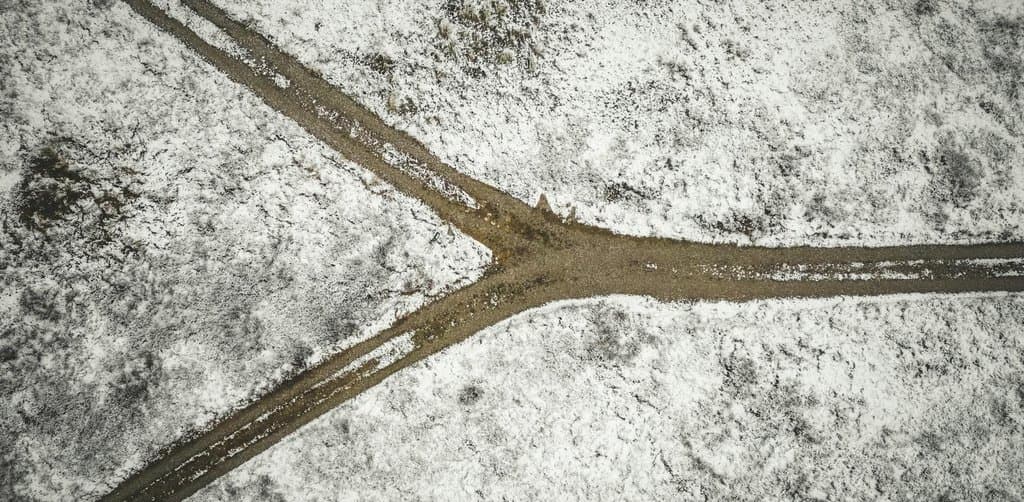 An aerial overhead view of a snow-dusted muddy brown country road where a single dirt path splits into a Y-shape with two diverging branches running off the bottom edges of the frame across a flat field of light snow-covered grassland and bare scattered shrub vegetation with subtle tire ruts visible in the muddy soil under directly overhead winter sunlight, the literal directional-decision metaphor for the route choice every participant makes at an employee stock ownership plan cash out election