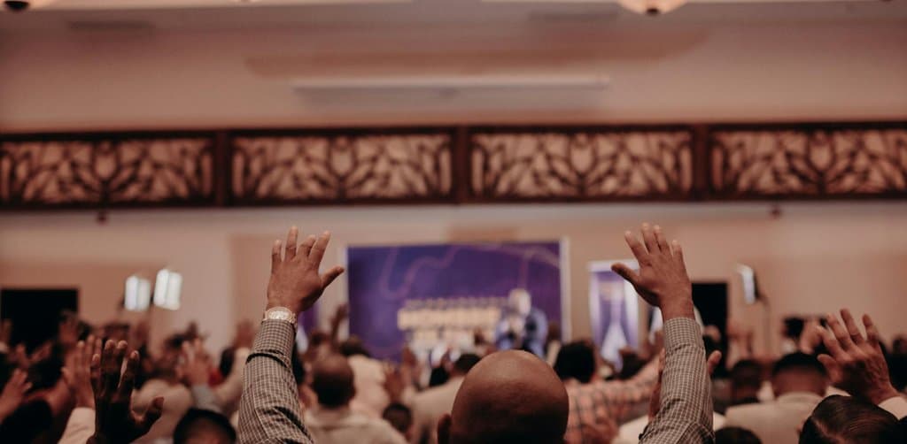 A warm-lit ballroom or banquet hall scene shown from behind the audience showing several rows of dark-suited mostly male attendees with arms raised toward a brightly lit stage with a white-toned screen and a single speaker silhouetted against blue and red stage lighting under three pendant globe chandeliers visible at the top of the frame against ornate wood-paneled lattice wall trim, the literal raised-hand questions metaphor for the broad participant interest in employee stock ownership plan faqs across every ESOP-owned company