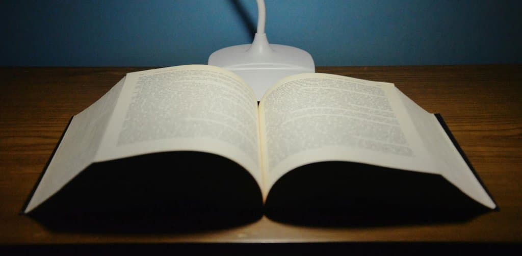 A small black hardcover book lying open flat on a dark wood-grain plank desk with both pages fanned out under a single white articulating modern desk lamp positioned directly overhead casting bright light onto the open pages with the lamp neck visible rising above the book against a deep teal-blue plaster wall background, the literal reference-document metaphor for the role of the plan document and the employee stock ownership plan faqs document in answering every participant question about plan rules