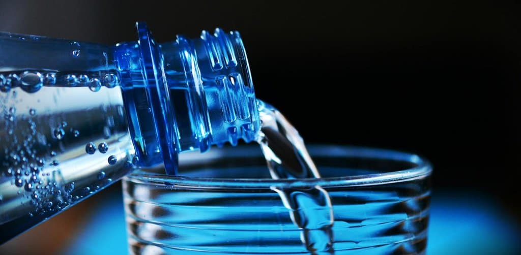 A close-up product shot of a clear blue plastic water bottle tipped on its side and pouring a steady stream of water into the top of a clear glass tumbler with horizontal ringed glass texture against a black background with cool blue ambient lighting and visible air bubbles inside the empty bottle, the literal flow metaphor for moving balance from an esop into a new qualified account through a clean employee stock ownership plan rollover
