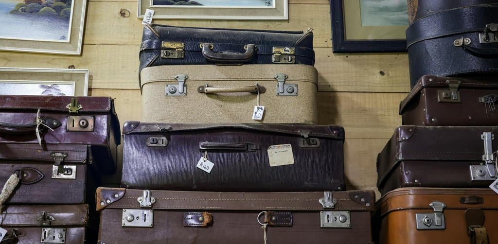 A vertical stack of seven vintage hard-shell travel suitcases of different sizes and dark earthtone colors black brown rust and tan with metal latch hardware aged paper price tags handwritten with prices like R120 stacked on a wood plank floor against a wood-paneled wall with three framed pastel landscape paintings of distant mountains above the luggage stack, the careful documented transfer metaphor for the structured employee stock ownership plan rollover paperwork an esop trustee processes each year