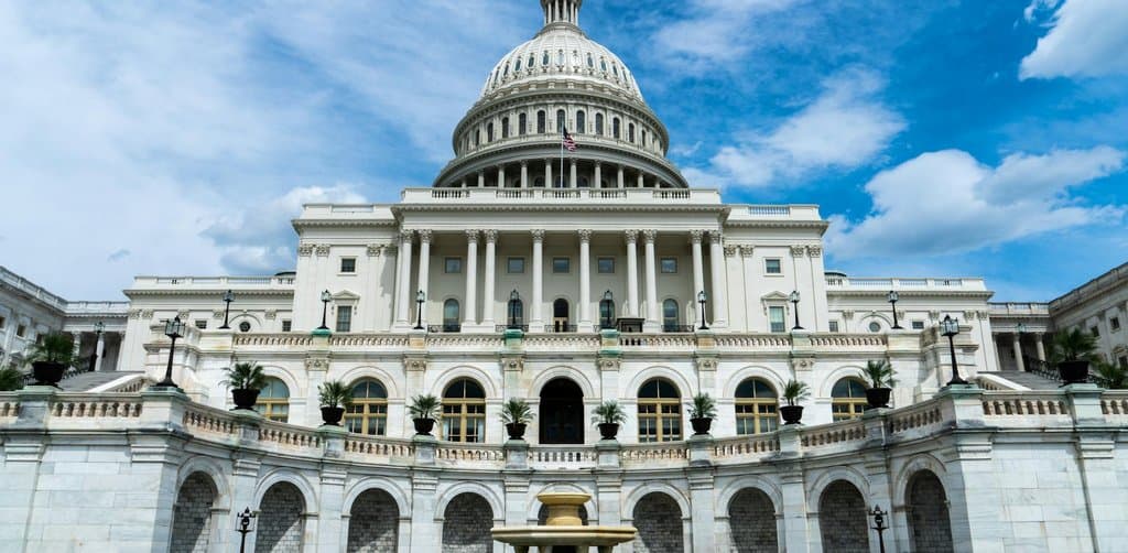 A wide front-facing view of the U.S. Capitol building in Washington DC with the iconic white sandstone dome topped with the Statue of Freedom rising above the central porticoed colonnade and clean white stone wings extending left and right with a tiered fountain and circular drive in the lower foreground under a bright blue sky with scattered cumulus clouds and a US flag visible to the left of the dome, the literal federal-law and legislative-rules metaphor for the IRC tax code provisions behind employee stock ownership plan taxation