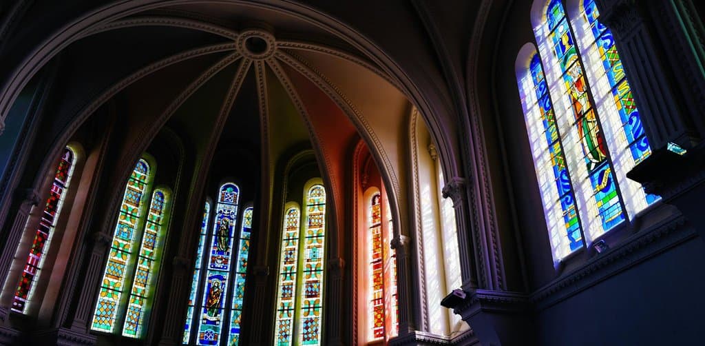 A vaulted cathedral interior with tall narrow Gothic stained glass windows in blues reds greens and yellows casting colored shafts of light across the columned nave with a ribbed white plaster ceiling overhead and a single saint figure window glowing brightly on the right transept wall, the multi-layered structural complexity that mirrors the layered legal trust valuation and tax frameworks behind every esop