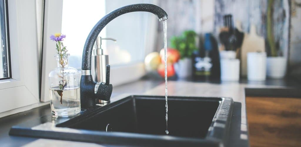 A bright modern kitchen sink corner scene showing a tall black faucet with chrome accents pouring a clear stream of water into the black granite sink basin beside a windowsill holding a small clear glass bottle vase with a single purple aster flower and a chrome soap dispenser with various countertop accessories blurred in the background, the literal flowing tap metaphor for how esop dividends drip from the corporate cash flow into participant accounts each year on a steady cadence