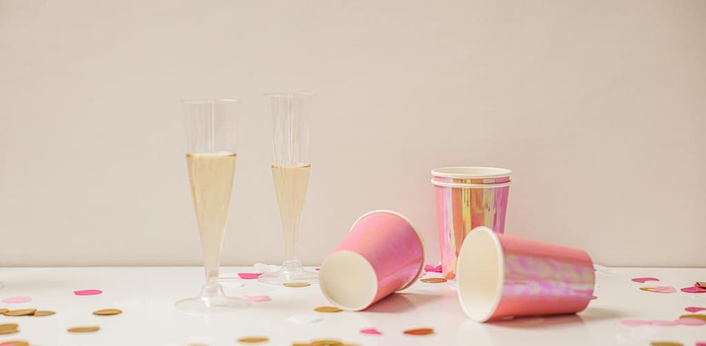 A bright clean product still life on a white tabletop showing two clear plastic champagne flutes filled with golden sparkling wine standing upright in the center beside three iridescent pink-and-gold paper drinking cups one upright and two tipped over with scattered pink and gold round confetti dots across the white surface against a soft pale cream background, the celebration moment metaphor for the immediate cash distribution participants receive at the esop payout when company is sold