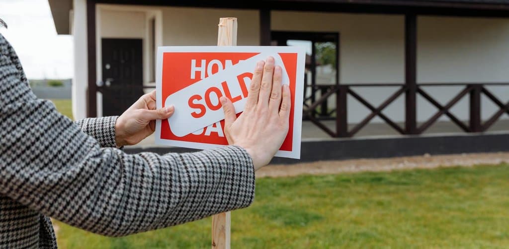 A close-up profile view of two hands belonging to a person in a black-and-white houndstooth pattern coat applying a diagonal white sold strip sticker across a red For Sale yard sign mounted on a wooden post stake driven into green grass in front of a residential white-and-brown ranch-style house with covered porch and gabled black roof in soft outdoor daylight, the literal sale-closure metaphor for the moment of esop payout when company is sold and the trust receives the transaction proceeds
