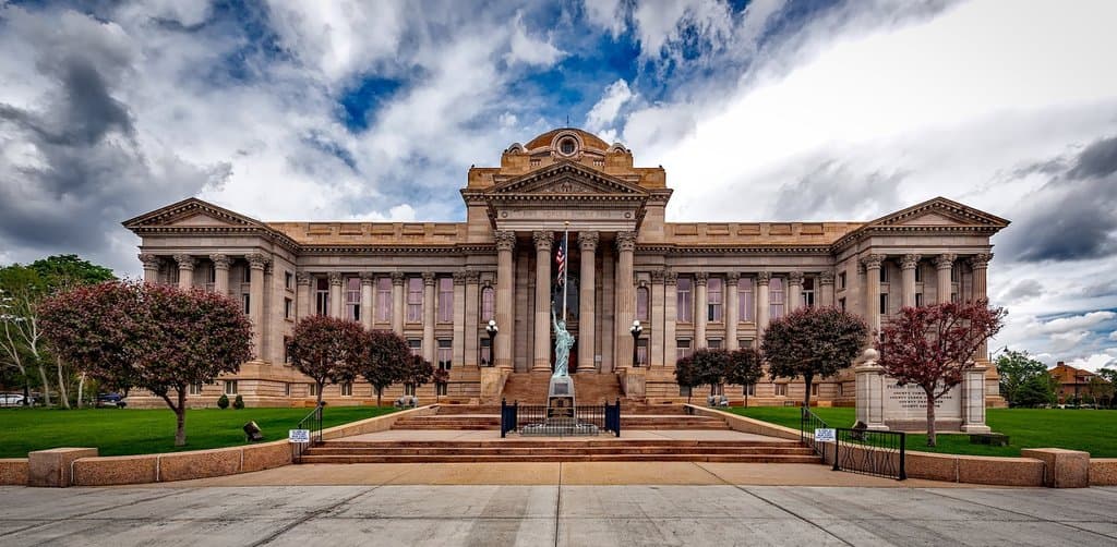 A grand neoclassical sandstone county courthouse with a wide columned portico tall white Corinthian columns a copper-domed central pavilion an American flag on a tall pole at the center steps a green bronze statue at the base of the steps two ornate sandstone wing pavilions and a dramatic cloudy sky overhead with two ornamental red-leaved trees framing the walkway in the foreground, the formal fiduciary court oversight imagery behind the esop trustee role under ERISA
