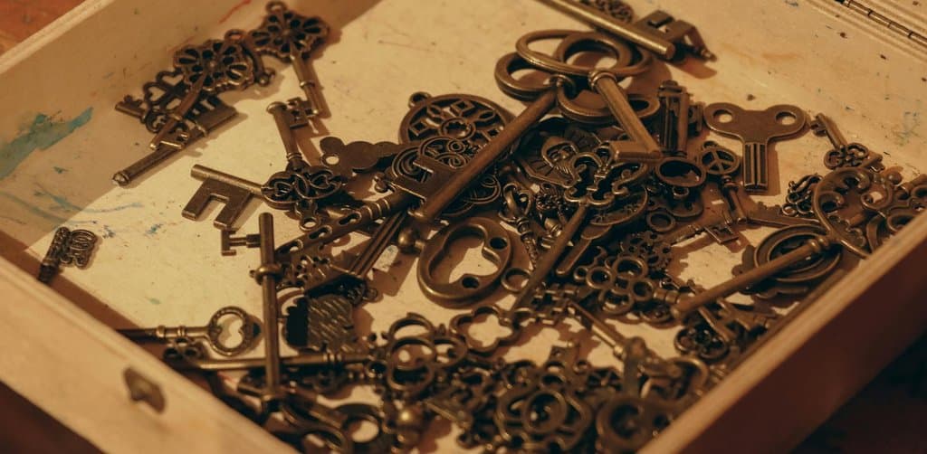 A shallow square wooden box on a paint-spattered workbench overflowing with dozens of ornate antique brass skeleton keys of various sizes and styles in warm directional lighting that catches the bronze patina of the metalwork and the deep shadows in the corners of the box, the trustee custody and stewardship metaphor for the esop trustee role of safeguarding plan shares on behalf of every participant