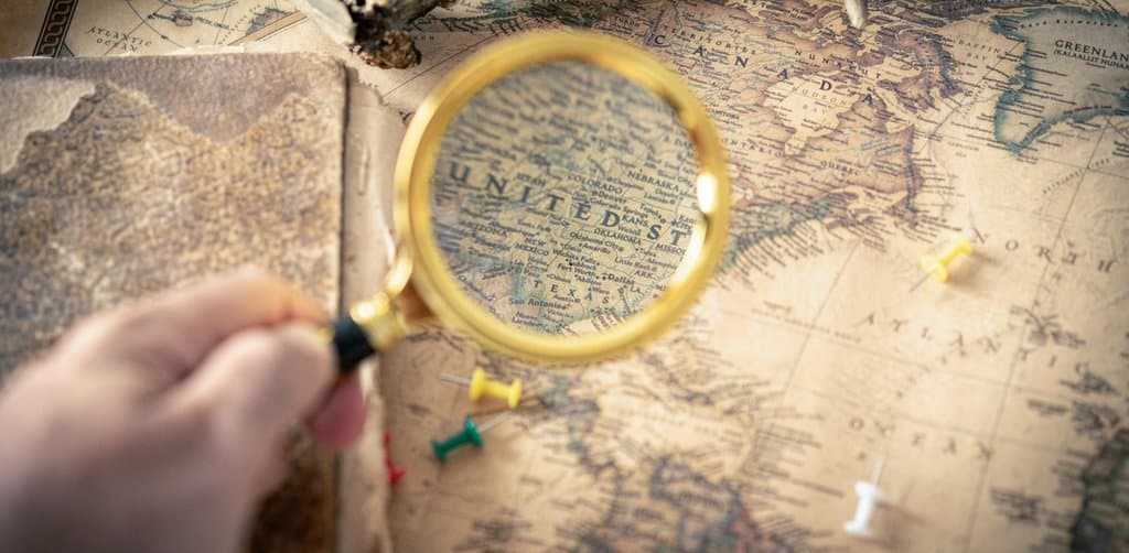 A golden brass magnifying glass with a black handle held in a hand over an antique parchment-toned world map showing the United States Canada Alaska and Mexico with tiny colored pushpins scattered across the surface and a piece of weathered wood at the top of the frame and an open antique notebook on the left edge, the close-examination metaphor for how an esop valuation appraiser scrutinizes every line in the financial statements