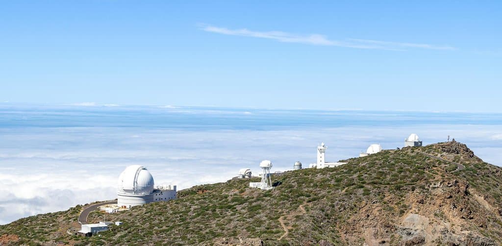 A long mountain ridge with multiple white-domed astronomical observatory buildings spaced along a rolling green-and-rust-colored summit ridge under a clear blue sky with a sea of white cloud cover stretched across the lower elevations and a distant blue horizon, the precision instrument metaphor for how an esop valuation looks at the company from multiple methodological angles simultaneously