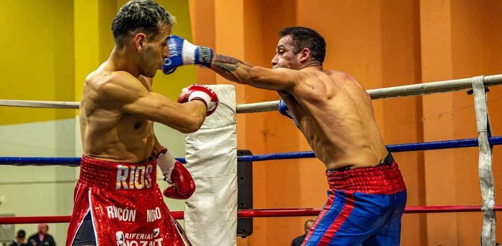 Two welterweight boxers face off in the corner of a brightly lit ring with the boxer on the left in red trunks reading RIOS slipping a glove punch from the boxer on the right in red-and-blue striped shorts who is throwing a straight right hand under an orange-yellow padded pillar with the metal ring ropes visible in the foreground, the head-to-head competition framing of the esop vs 401k comparison sellers and employees run through every year