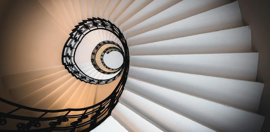 A direct overhead architectural shot of a tall narrow spiral wooden staircase with white-painted balustrades and dark wrought-iron handrail decorations winding down through multiple stories of a small foyer with a single warm pendant light bulb glowing at the center of the spiral well and afternoon shadows cast diagonally across the lower flights of stairs, the spiraling layered structural comparison metaphor for evaluating esop vs rsu equity awards across multiple time-bound steps of vesting and tax events