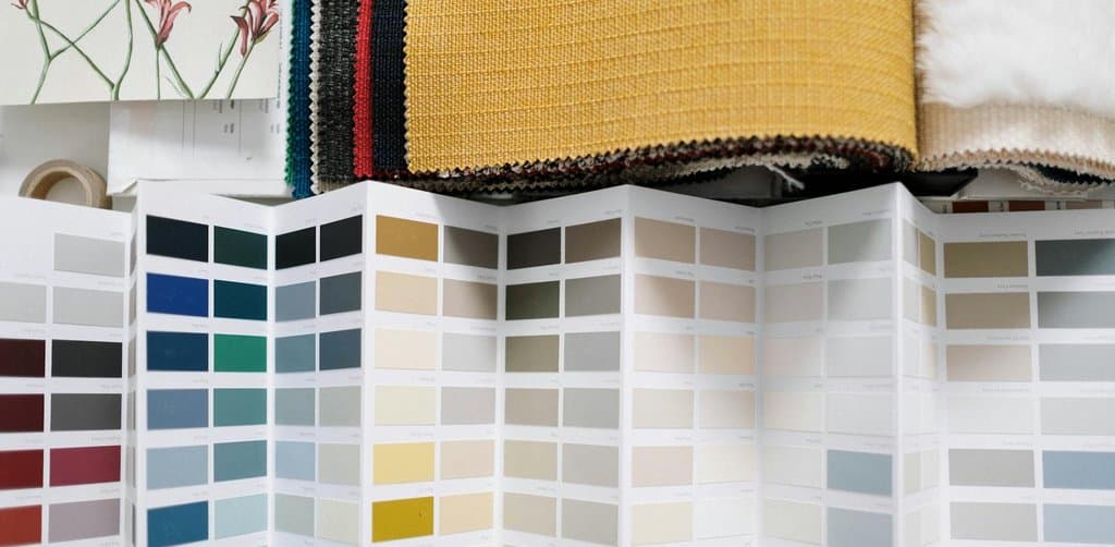 A close-up of a designer's material consultation tabletop showing a vertical fan of paint color swatch cards in dozens of muted earth tones whites greys creams blues and reds with fabric samples in yellow navy cream and red striped layered above the swatch fans and a small floral wallpaper sample on the left and a soft white fur sample on the right, the side-by-side feature comparison metaphor for evaluating esop vs rsu equity awards across multiple variables at once