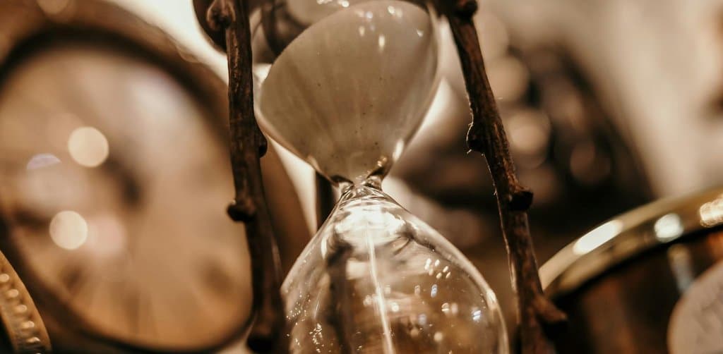 An intimate macro close-up of an antique brass-and-glass hourglass timer with a wrought-iron frame in the foreground showing fine white sand mid-flow falling from the upper bulb through the narrow waist into the lower bulb against a softly out-of-focus background of warm vintage brass clock faces and gear edges in warm light, the time-bound flow metaphor for esop withdrawal rules that allow access only after specific time thresholds and triggering events have passed
