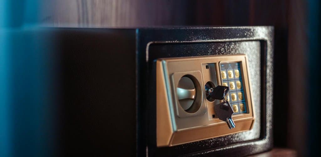 A close-up of a small black metal lockable home safe sitting on a dark wood shelf against a deep blue ambient wall with a gold-toned brass front panel and circular turn-dial handle on the left and a black backlit keypad with golden number keys on the right and a small black security key inserted with key ring hanging from the lock mechanism, the literal protected access control metaphor for the strict esop withdrawal rules that govern when participants can take cash out of their esop accounts