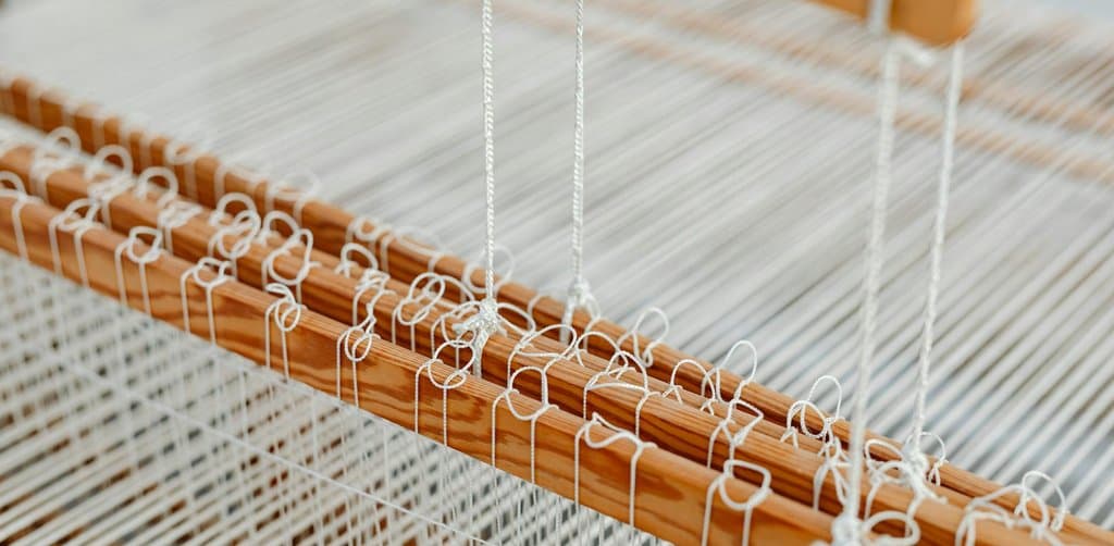 A horizontal wooden table loom in close-up showing tightly strung vertical white cotton warp threads passing through a pine wooden heddle frame with dozens of small white string loops dangling from the heddle bar and rows of long natural-fiber loops attached along the top wooden frame indicating different yarn shafts under bright natural daylight, the structured weaving allocation metaphor for how are esop shares allocated across plan participants on the strict frame of the IRS rules