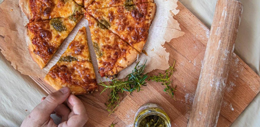 An overhead view on a wooden cutting board of a freshly baked round pizza already cut into eight wedge slices arranged in their original circular formation with melted cheese and pesto and a small jar of green herb sauce on the side a sprig of rosemary and the worn wood handle of an old rolling pin visible at the right edge as a left hand reaches in from the lower left to take a slice, the literal pro-rata division metaphor for how are esop shares allocated to each participant in proportion to their share of total payroll