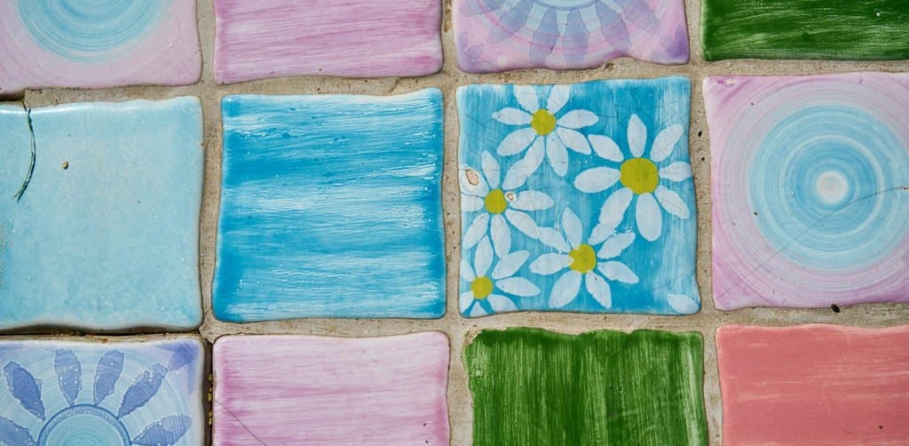 A close-up overhead view of nine hand-painted ceramic floor tiles arranged in a three-by-three grid with grout lines visible between them each tile a different solid color or simple pattern including pale pink turquoise white-and-yellow daisy flowers light green sky blue sunburst and a coral salmon pink the grid composition metaphor for how are esop shares allocated across participants in a defined allocation grid every plan year
