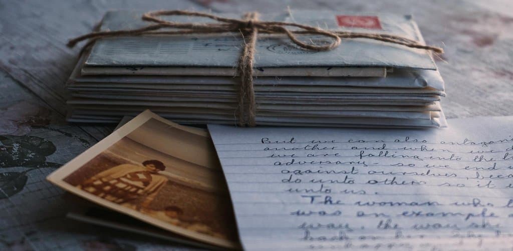 A stack of dozens of old creased handwritten letters and small white envelopes tied together with frayed jute twine in a hand-tied bow on a weathered wooden surface alongside an antique sepia-toned black-and-white photograph of a young woman seated on a wicker chair and a single open lined page of cursive handwritten letter content visible at the right edge, the careful documentation and patient mail-driven correspondence metaphor for how do i cash out my esop after i quit a job through the formal multi-year distribution mail flow