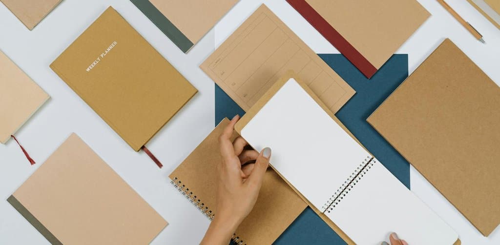 A wide flat-lay overhead view of dozens of small kraft-paper-covered weekly planner notebooks of different sizes and shapes spread across a white desktop surface with a single navy blue page beneath an open spiral-bound planner being held open by two female hands with manicured nails the open planner showing blank lined paper on the right and a kraft tab on the left, the careful documentation and scheduling metaphor for the structured timeline behind how do i cash out my esop after i quit a job