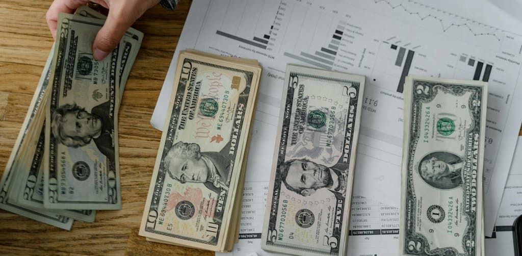 Overhead view of stacked twenty, ten, five and one dollar bills arranged on top of financial bar charts and reports, illustrating the lump-sum cash a note buyer pays after running discount-rate math on a seller-financed business note