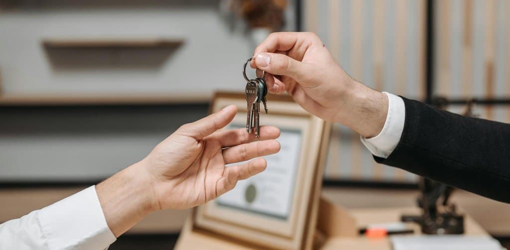 Suited professional handing a set of keys to a buyer across a desk with a framed certificate behind, the closing handover moment of a small business acquisition that funded with little to no buyer cash at the table