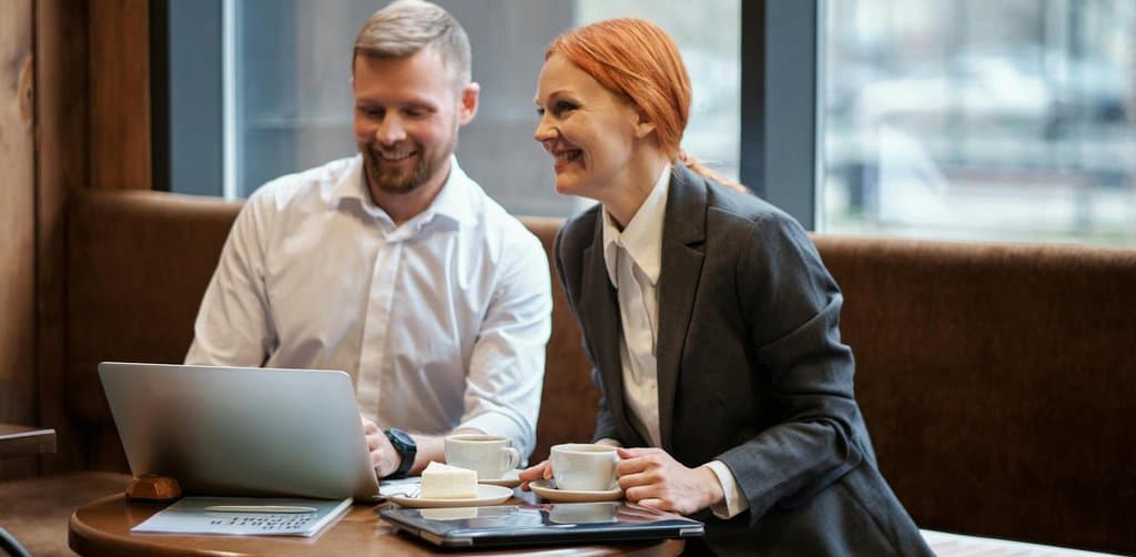 Two professionals seated at a coffee shop table with a laptop and coffee cups, the buyer-seller diligence conversation that typically precedes a no-money-down small business acquisition