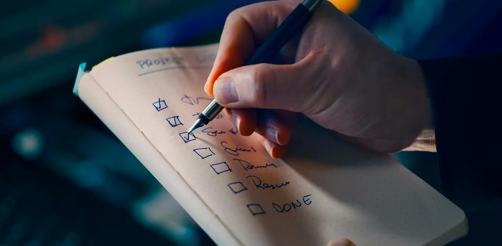 Hand using a fountain pen to check off items on a handwritten dot-grid notebook checklist with items already marked done, the working diligence list a buyer maintains while running every lien and litigation search before closing on a business