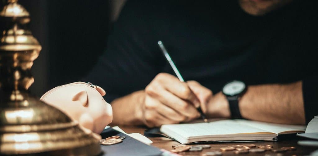 Man writing in a ledger beside a piggybank and coins, illustrating the payoff arithmetic borrowers run before learning how to remove a business lien