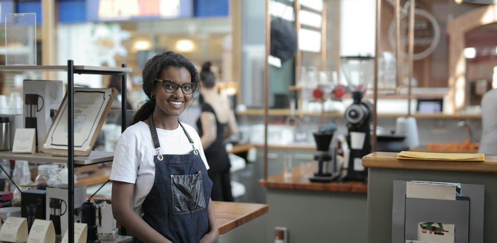 A smiling young woman with curly hair wearing a denim apron and white t-shirt standing in her cafe shop with espresso machines and merchandise behind her, the kind of small business owner who benefits from the new SBA loans 2025 program updates