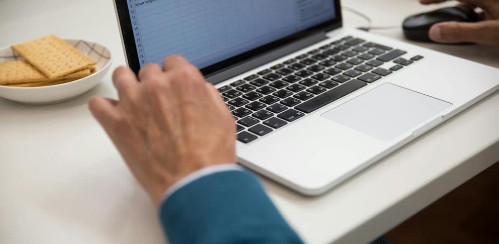 A close-up of a mature man's hands on the keyboard of an open silver laptop displaying a spreadsheet of numbered rows containing names like Pedro, Jose, Francisco, and Celina with a saucer of crackers and a black mouse on the white desk beside him, the careful daily ledger work that defines note servicing best practices