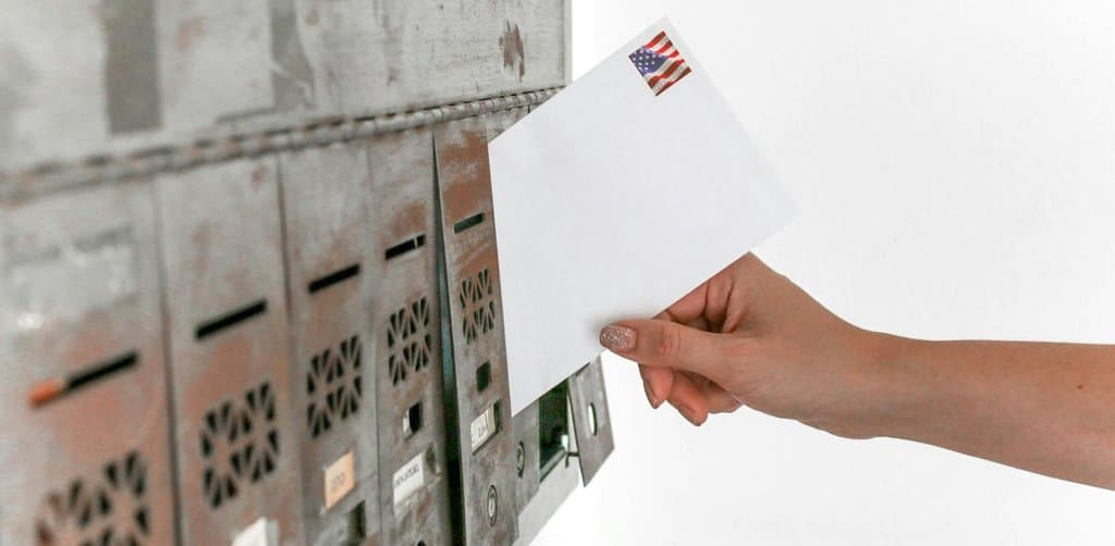 A woman's hand with manicured nails holding a sealed white envelope with a small American flag postage stamp affixed to the upper right corner positioned in front of a row of weathered metal mailbox slots with peeling paint and small ventilation grates, the formal delivery channel that preserves proof of every late-payment notice in a servicing file