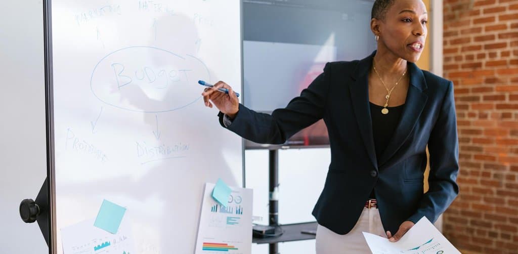 A woman in a dark blazer presenting a Budget flow diagram on a whiteboard with sticky notes and printed bar charts, the founder pitching the use-of-proceeds story that anchors any serious round of raising capital