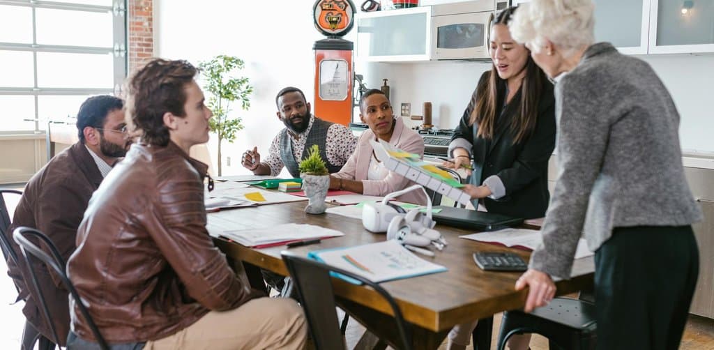 A group of professionals seated around a wooden table in a loft workspace reviewing printed financial charts and a colorful pitch document, the kind of multi-party founder and investor meeting that anchors a typical capital raise