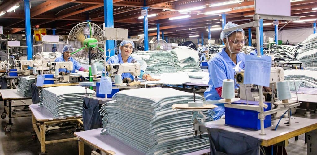 Three workers in blue uniforms at sewing stations inside an industrial garment factory with stacks of folded fabric on each work station, the kind of light manufacturing operation that frequently uses SBA 5(04) loans to fund equipment and facility purchases