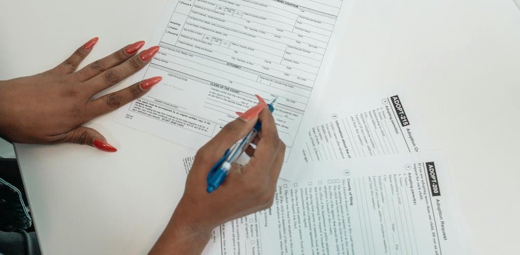 Hands with red-painted fingernails filling out a stack of multi-page government forms with a blue pen on a white desk, the application paperwork that defines the SBA 5(04) loan diligence and qualification process