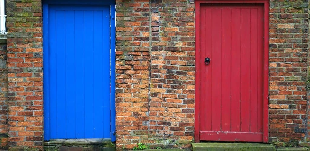 A weathered red painted door and a bright blue painted door set side by side in a worn red-brick exterior wall on a sidewalk, the simple two-program choice that defines SBA 7(a) vs 504 in acquisitions decisions