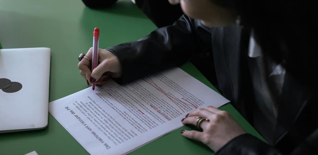 A close-up of a woman in a black leather jacket leaning over a green office desk and editing a French-language printed manuscript with a red ballpoint pen, the kind of policy revision work that produces ongoing SBA changes
