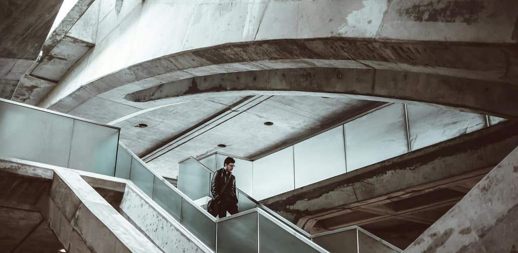 A man in a dark jacket walking across a suspended pedestrian bridge inside a dramatic curved concrete and steel modernist building with high arched ceilings, an architectural representation of moving through the structural transitions of SBA changes