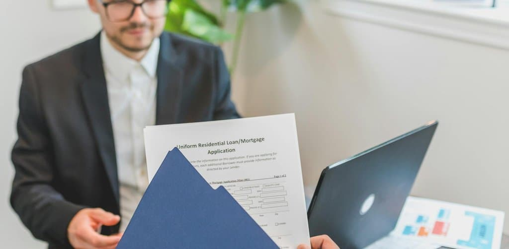 A close-up of a Uniform Residential Loan and Mortgage Application paperwork being held up at a desk with a blurred suited man in a black blazer behind it, the kind of formal application that captures the SBA equity injection requirements 2025 documentation