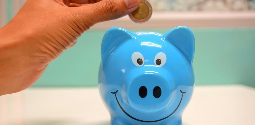 A hand placing a coin into a blue ceramic piggy bank with painted smile and eyes on a white surface against a teal background, illustrating the personal savings discipline that funds the buyer cash portion of SBA equity injection requirements 2025