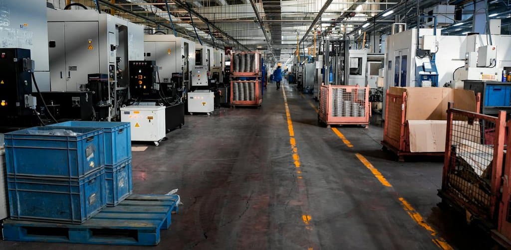 A wide overhead view of an industrial factory floor with rows of CNC machines along both sides, blue and red pallet bins, and a yellow dividing line running down the center aisle, the kind of operating business that frequently changes hands through SBA loan assumption rules