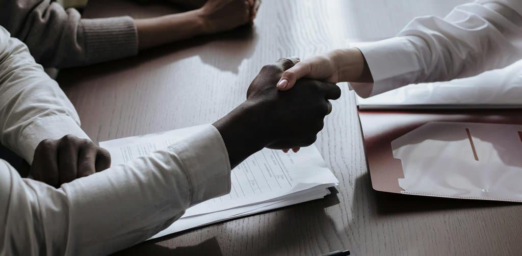 Two professionals shaking hands across a dark wooden desk with stacks of paperwork and a red folder, sealing the formal closing moment of an SBA loan assumption between original borrower and assuming buyer