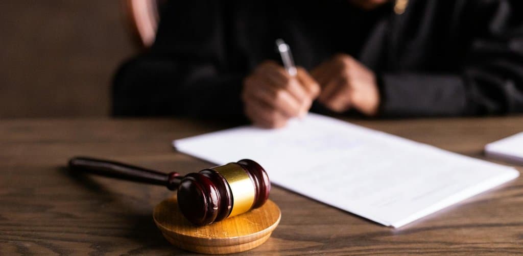 A wooden gavel resting on its sound block at the front of a wooden desk with a focused woman in a black blazer writing notes on a legal document in the background, illustrating the courtroom and legal-process side of an SBA loan default