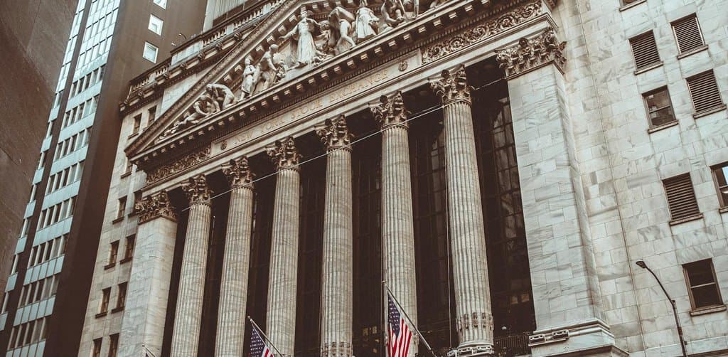 A street-level view of the New York Stock Exchange building facade with classical columns and three large United States flags hanging across the front, the secondary market environment in which the SBA loan lender secondary market operates